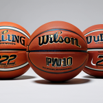 A professional studio photograph showcasing a diverse collection of basketballs, emphasizing material innovation. In the foreground, a durable, textured rubber outdoor basketball designed for rough surfaces. Next to it, a sleek, premium composite leather indoor basketball, highlighting its superior grip and refined surface. A third, subtly visible ball in the background features advanced micro-textures or a sustainable material label, hinting at future innovations. The composition is clean and minimalist, with soft, even lighting. High-resolution, crisp focus, vibrant sports aesthetic, safe for work, appropriate content, fully clothed, family-friendly, perfect anatomy, correct proportions, natural composition.